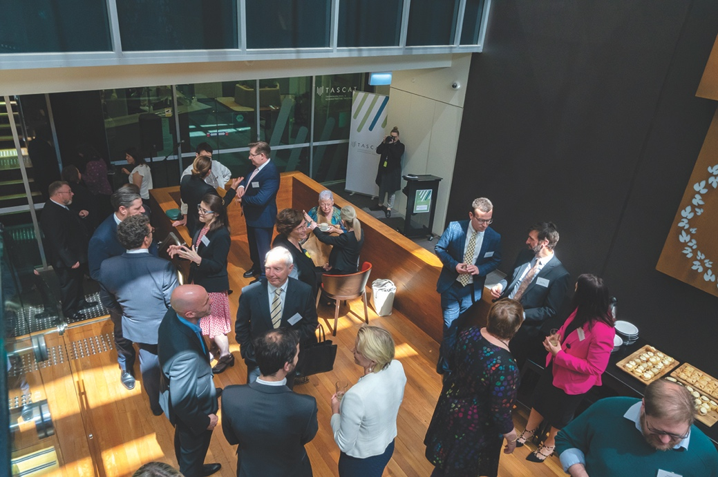 Group of around twenty people in formal business clothes at a function for the opening of TASCAT, taken from above, on a timber floor with a glass wall at the back of the room