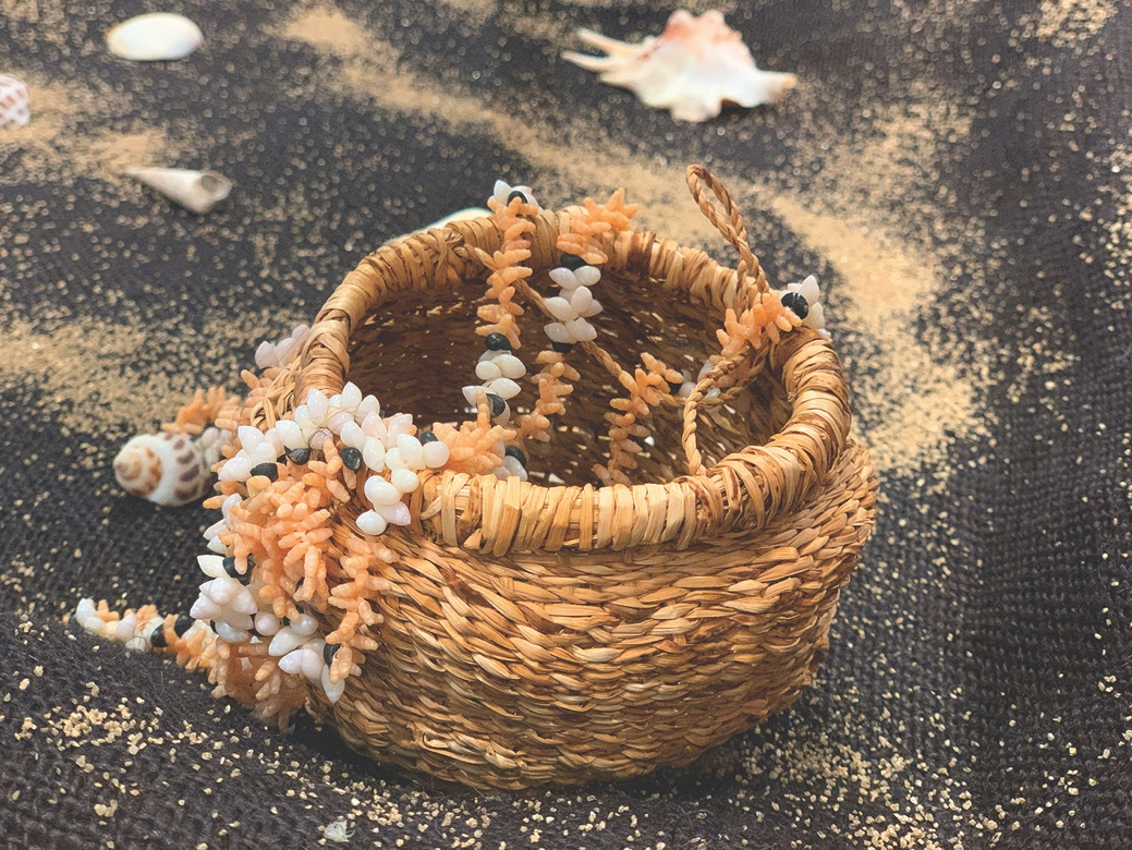A woven basket containing a white shell necklace, on a dark cloth covered in sand, with shells scattered around