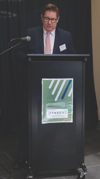 Malcolm Schivens, President of TASCAt, giving a speech at a podium. He has medium-brown hair and is wearing a dark blue suit, white shirt and a red tie with small white squares.