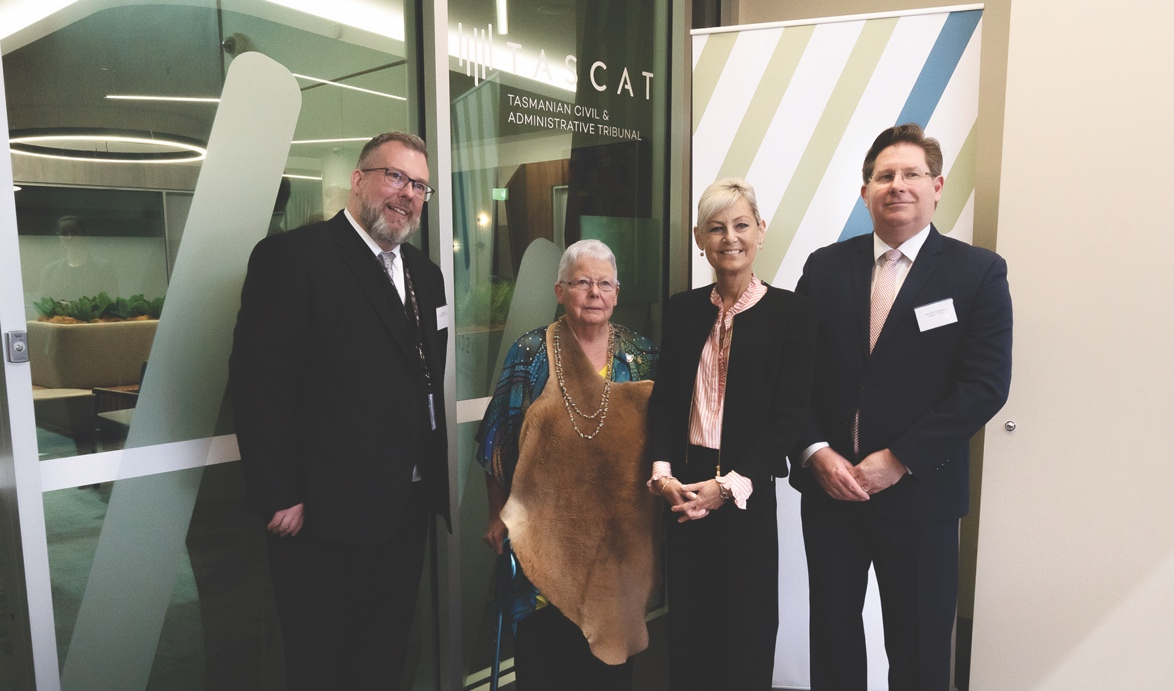 Four people standing in a row in front of a glass door at the entrance to the TASCAT offices. The two mean are at each end of the group and are dressed in business suits. Minister Archer is wearing a dark  suit and a salmon-and-white striped shirt. Auntie Brenda Hodge is wearing a wallaby skin over a blue shirt with an Aboriginal print.