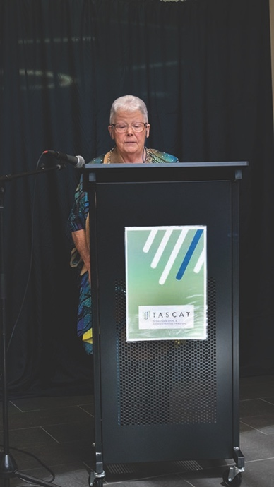 Auntie Brenda Hodge gives the Welcome to Country standing behind a podium. She has short grey hair and is wearing a blue shirt with an Aboriginal print